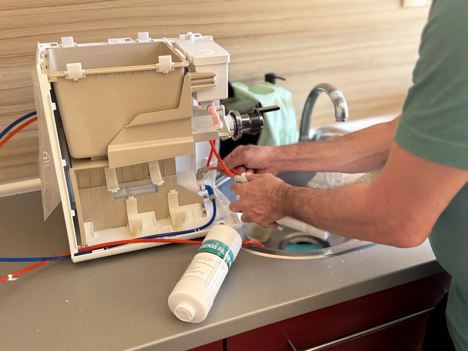 A technician's hands connect hose fittings on a disassembled appliance at a kitchen sink.